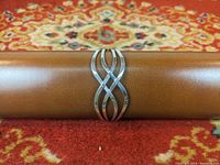 Full view of the front side of the silver cuff bracelet showing the intertwined open design and polished finish on a brown cylindrical display with a colorful patterned background.