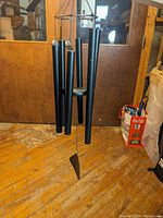 Large black metal wind chime with six hanging tubes of varying lengths, shown hanging indoors against wooden door background on wooden floor.