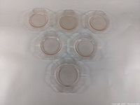 Top view of six pink depression glass bread and butter plates arranged in two rows of three on a white surface showing their translucent pink color and scalloped edges.