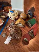 Various hand painted birdhouses and a metal wind chime arranged on a wooden floor, showing the variety of shapes, sizes, and colors including a natural twig house and painted wooden houses.