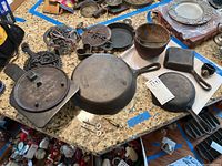 Wide angle photo of the full lot on kitchen counter including multiple cast iron pans, flue cover, pail, heart shaped bowl, skeleton keys, lock and trivets.
