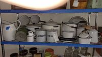 Full view of white enamel ware kitchen items on metal shelves including large pitcher, strainers, labeled canisters, pots, and cups