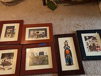 Six framed art prints arranged together on floor, showing variety of folk art scenes and figures.