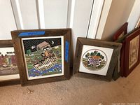 Overall view of several framed folk art prints with rustic wood frames leaning against a wall on carpeted floor
