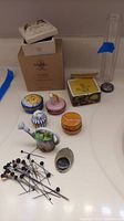 Photo of various pill boxes, including Limoges watering can shaped box, Royal Copenhagen box in brown container, Sun-themed round box, and glass vase in background, with decorative hat pins in front