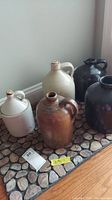 Group view of all 5 ceramic jugs on a stone-patterned mat, showing varying sizes and colors with some wear.