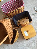Photo showing a group of baskets including a medium basket with dark blue liner, small oval basket with wooden lid, small square basket with wooden lid, and other baskets on carpet