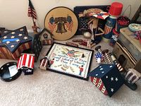 Wide view showing various patriotic decorations including star boxes, birdhouses, framed embroidery, painted tin, wooden figure, and thermos.