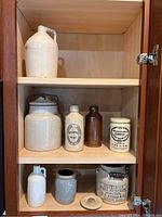Eight piece stoneware collection arranged in wooden cabinet shelving including crocks, jugs, and labeled containers