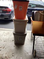 Stack of seven assorted plastic storage bins in orange and light gray colors with lids in garage setting, showing overall lot.