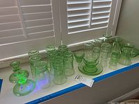 Wide shot of 28 green translucent uranium glass pieces including jars, cups, and candlesticks on a windowsill under UV light showing fluorescence.