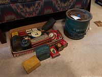 Overall view of vintage tins laid in a box and the green Emsco minnow bucket on the floor.