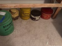 Photo showing six vintage tins lined up under a wooden bench: Stehman's Potato Chips green tin, two Charles Cookies tins (large and small), Tommy Dale Potato Chips tin, Miller Butter Brand tin, and Sturgis Pretzel Company red tin.