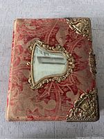 Front view of antique photo album showing red patterned fabric cover, decorative metal corners, metal framed window inset, and clasp lock.