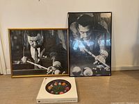 Two framed black and white photos of men playing pool and a round billiards-themed clock on floor against wall