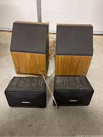 Front and top view of 4 Bose speakers including 2 wooden cabinet and 2 black plastic speakers with speaker wire on garage floor