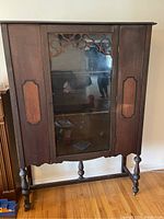 Front view of vintage wooden china cabinet showing central glass door with decorative fretwork, side wood panels, and carved legs.