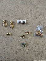 Full lot overview of various clip-on earrings on a beige surface, showing multiple pairs including gold-tone, floral, and small stud styles.