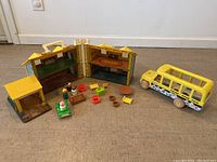 Foldable dollhouse open flat with multiple small furniture pieces arranged in front and yellow bus partially visible on right side.