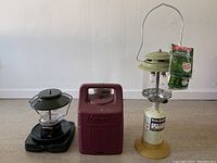 Two propane lanterns and a maroon carrying case arranged on floor against wall