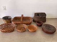 Eight assorted wicker baskets arranged on floor. Various shapes and sizes including oval, round, rectangular, and a basket with a damaged lid.