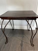 Front view of the custom side table showing the dark wood tabletop and curved iron legs against a white wall and wood floor.