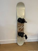 Full view of the K2 men's snowboard standing on its tail against a white wall, with attached FLOW M9 bindings visible.