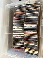 Top view of a plastic bin stacked with numerous music CDs in jewel cases, showing front and spine labels of various albums from the 80s and 90s.