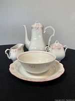 Full view of the 5-piece porcelain mini coffee set on a black surface showing coffee pot, creamer, sugar bowl with lid, cereal bowl, and dessert plate decorated with pink ribbon bows.
