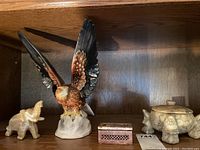 Four decorative items displayed on wooden shelf including alabaster elephant, ceramic falcon, stone box, and marble bowl with lid