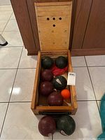 Wooden case open showing four bocce balls (two maroon, two green), red cloth, orange and white small balls inside, and lid with ventilation holes.