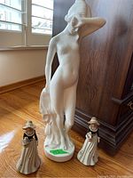 White nude ceramic figure standing with two smaller ceramic maiden figurines in front on wood floor near wooden furniture and window.