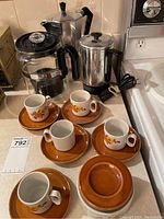 Photo showing four Sebring Porceliana espresso cups with floral patterns on brown saucers, one extra saucer, an Empire electric percolator, and Bialetti Moka Express espresso maker on kitchen counter.