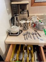 General Electric Coffeematic coffee maker with clear glass pot and black handle, stainless steel stovetop coffeemaker, white teapot, hand painted creamer and sugar bowl set, silver spoon, plus various cups and saucers with teaspoons on countertop.