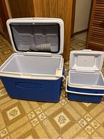 Large Rubbermaid cooler and two small Rubbermaid coolers with white lids inside on floor with wood panel wall background.