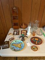 Overview of all items on table including Avon mallard after shave bottle, hobnail milk glass vase, clear glass vases, wooden mail holder, ceramic travel plates, small ceramic teapot, framed art and hanging chimes.
