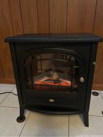Black Dimplex electric heater fireplace shown from front right angle on tiled floor against wood panel wall, plugged in power cord visible