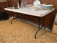 Full view of folding table showing plastic top and black metal folding legs, set up indoors on brown carpet in front of wood-paneled wall.