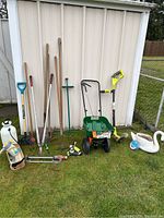 Full view of garden tools and equipment arranged outside shed on grass, includes hand tools, spreader, weed eater, garden decorations, and sprayers