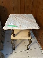 Square marble top end table with veining and scalloped edges, supported by four ornately detailed metal legs and a lower marble shelf, placed on white tile floor against wood panel wall.