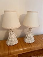 Pair of bedside lamps with lady figurine bases and cream fabric shades, shown side by side on wooden surface.