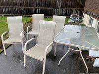 Photo of the full patio set showing four beige colored mesh sling chairs and rectangular glass table on wet patio surface