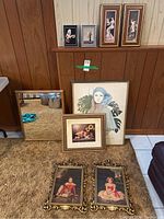 Display of all framed prints and wood framed mirror on carpeted floor against wall showing the variety of sizes and styles, including large portrait print, floral print, two child portraits with gold frames, and mirror.