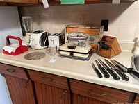 Wide shot showing all lot items on kitchen counter including toasters, waffle grill, blender, knives, napkin holder, glassware