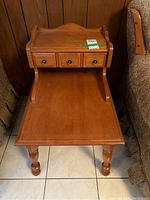 Maple side table front view showing three small drawers on upper shelf, curved side edges, and turned legs on tiled floor between couches.