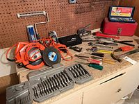 Photo showing the arrangement of tools on workbench, including Orange Black & Decker circular saw, various hand tools, drill bit sets, and small toolboxes.