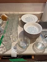 Photo showing stacks of white ironstone dinner plates and soup bowls with leaf pattern, Pyrex clear glass bowls, and multiple clear water glasses on kitchen counter