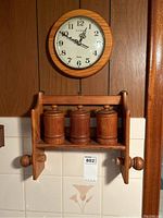 Front view of wooden quartz clock above wooden wall-mounted shelf with three canisters and paper towel holder.