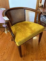 Wooden arm chair with curved armrests and woven cane backrest, upholstered with a mustard yellow cushion, positioned on hardwood flooring near a pink lamp and doorway.