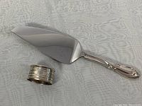 Photo of a 10.5 inch cake cutter with ornate sterling silver handle alongside a silver napkin ring with monogram initials H B.
